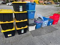 Photo showing 8 large black bins with yellow lids stacked in two columns, various other blue, gray, white, and clear bins, some lids missing; taken outside on pavement.