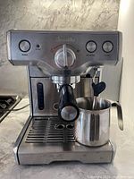 Front view of stainless steel Breville espresso maker with milk jug positioned on drip tray, machine turned on with illuminated power button.