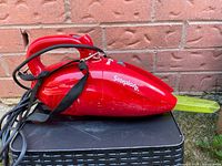 Side profile photo showing the full red handheld vacuum with yellow nozzle and attached cord and strap, placed on a black surface against a brick wall.