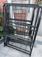 Folded black metal double bed frame shown from side angle outside near brick wall and plants.