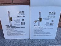 Two original product boxes of Home Decorators Collection Marta 2-Light Vanity Fixtures with gold finish and clear ribbed glass.