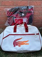 Two gym bags stacked: white Lacoste bag with red crocodile logo in front, black and red GoodLife Fitness bag in original plastic wrap behind
