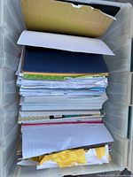 Top view of plastic storage bin filled with assorted scrapbook and craft papers, showing stacked sheets, some colored and patterned papers, and torn yellow cardboard piece.