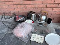 Image showing toaster, kettle, cast iron skillet, lemon squeezer, pink vase wrapped in bubble wrap, plates and wire basket on ground against brick wall.