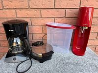 Photo showing Mr. Coffee coffee maker, Oster waffle maker, SodaStream soda maker, and Rubbermaid storage container on table against a brick wall