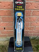 Photo showing the original packaging box of Optex T165 Tripod against a brick wall with the tripod visible in folded form.