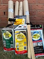 Photo showing 3 rolls of masking paper, 3 plastic drop sheets in different colored packaging, paint edge trimmer and wooden paint stirrer, all painting related items.