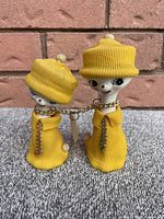 Front view of both dog figurines wearing yellow sweaters and hats, connected by a gold chain.