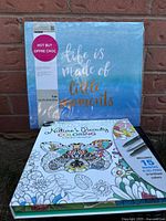Front cover of the scrapbook album with 'Life is made of little moments' text, wrapped in plastic packaging, alongside the Nature's Beauty coloring kit box with butterfly and flowers on the cover.