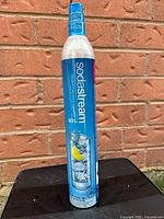 Photo of a new, sealed SodaStream 60L CO2 cylinder with blue branding and a lemon water splash graphic on the label against a brick wall background.