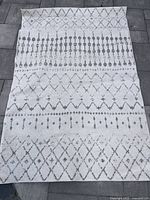 Front view of the rug laid out on a stone surface showing the geometric pattern in cream white and grey with diamond shapes and dotted lines.