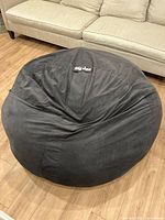 Black XL Big Joe bean bag chair photographed centered on wooden floor with beige couch behind it.