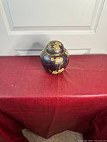 Front and side views of a solid brass decorative lidded jar or urn with gold floral accents on a dark background, placed on a red textured surface, 16 cm height approximately.