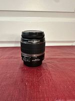 Front view of Canon EFS 18-55mm f/3.5-5.6 IS zoom lens standing upright on red surface.