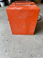 View of the heater from the back showing the solid metal housing and handle on top, orange painted with signs of use and wear.