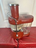 Front and side views of a red Fusion electric juicer with clear pulp container and silver locking arm, placed on a red cloth.