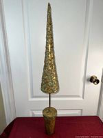 Full view of tall, narrow conical Christmas tree sculpture with ornate raised pattern and patina finish on decorative cylindrical base
