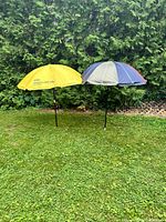 Two beach umbrellas set up outside on grass against a hedge background. One umbrella is yellow with black printed text, the other umbrella has three colored panels (blue, gray, red).