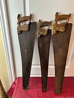 Three antique hand saws hanging vertically showing wood handles and steel blades with patina and rust spots.