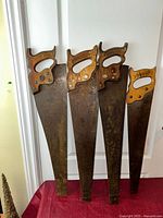 Four antique hand saws with wood handles and branded medallions, showing full length of blades and decorative handles.