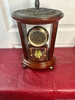 Front view of the wooden mantel clock with gold-tone round dial visible inside an oval wood and glass case with a decorative top knob.