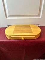 Front view of oval wooden cheese board with pull-out drawer closed, showing natural wood grain, drawer knob, and inlaid rectangular section