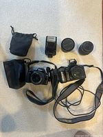 Overview photo showing two Nikon DSLR cameras, two lenses, a Nikon flash unit, and two black drawstring pouches laid out on a beige surface.