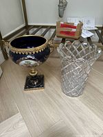 Overall view of the two vases, showing the porcelain urn on the left and clear glass vase on the right, displayed on floor with background furniture.