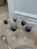 Photo showing five clear wine glasses with distinctive blue and green bubble decoration near the base of the bowl, arranged on a wooden floor.