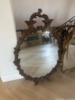 Antique oval mirror with intricate brown carved wooden frame, showing detailed scroll and leaf design and reflective glass.