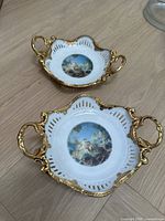 Two white porcelain decorative bowls with gold gilded handles and rims, featuring a classical painting scene in the center