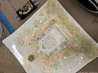 Top view of the decorative Judaica plaque showing Hebrew text, floral patterns, and small metallic dreidels attached around the text, with a metal flower ornament.