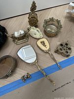 Assorted antique decorative items including hand mirrors, teapot, perfume bottle, trinket boxes, and decorative bowls in gold and silver tones on wooden floor.