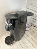 Front and side view of the black Nespresso coffee machine showing pod compartment, drip tray, and water reservoir.