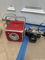 Photo showing Tekniq portable radio and Pentax SLR film camera side by side on floor.