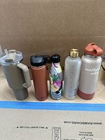 Front view of six reusable cups and bottles in varying colors and styles on cardboard background