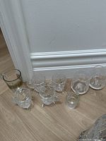Nine small glass teacups arranged on floor near wall showing variety of styles including cut glass and plain glass.