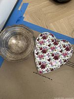 Two trays on floor: a round silver-tone metal tray with pierced sides and a heart-shaped ceramic floral tray.