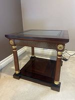 Front-side view of square side table showing wood frame, glass top, Greek key border, gold rosettes, and column supports.