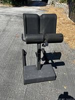 Photo showing the entire black back decompression device with foam pads and cylindrical roller on a metal base, placed outdoors on pavement.