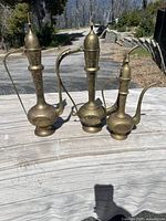 Three engraved brass urns displayed outdoors on a wooden surface under natural light, showing their shapes and engravings.