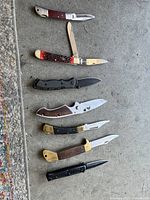 All seven pocket knives arranged vertically on concrete surface showing variety in handle styles and blade shapes.
