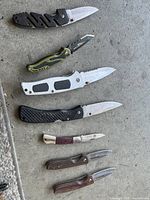 Top view showing six assorted pocket knives with different handles and blade styles placed on ground surface.