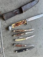 Six pocket cutters displayed on concrete floor including one large fixed blade knife with leather sheath and five smaller folding knives with various handles and blade conditions.