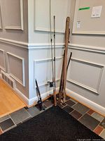 Seven antique fishing rods leaning against a wall on tiled floor, including wooden and composite rods with some covered in canvas sleeves.