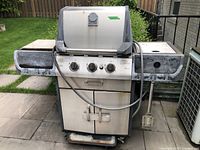 Front view of the Life at Home gas barbecue showing three control knobs, hose attached, and two side shelves with paint wear.