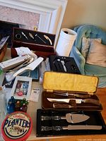 Photo showing carving sets in wooden cases with knives and utensils, a wine opener set in black case, silver serving utensils, Planters cocktail tin, and decorative bottles nearby.