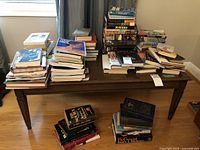 Wide view of multiple stacks of books including hardcover and paperback on a wooden table and floor.