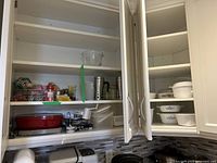 Wide shot of two kitchen cabinets with shelves containing Corning Ware dishes of various patterns, plastic containers, travel mugs, and kitchen accessories including a cheese grater and salt cellar.