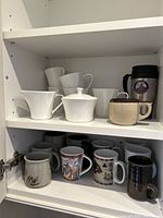 Photo of cabinet shelves holding various ceramic and pottery mugs, white creamer and sugar bowl, travel mug.